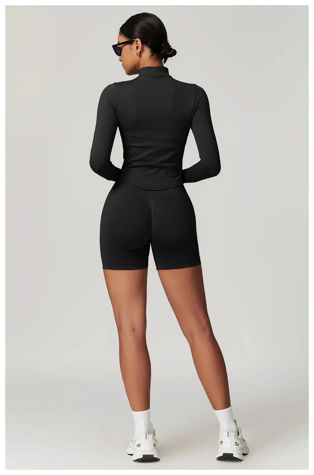 A woman stands facing away in a fitted Mirèlle Workout Jacket with Zip & Quick Dry, matching black biker shorts, white socks and sneakers, sunglasses, and her hair in a low bun, set against a light gray background.