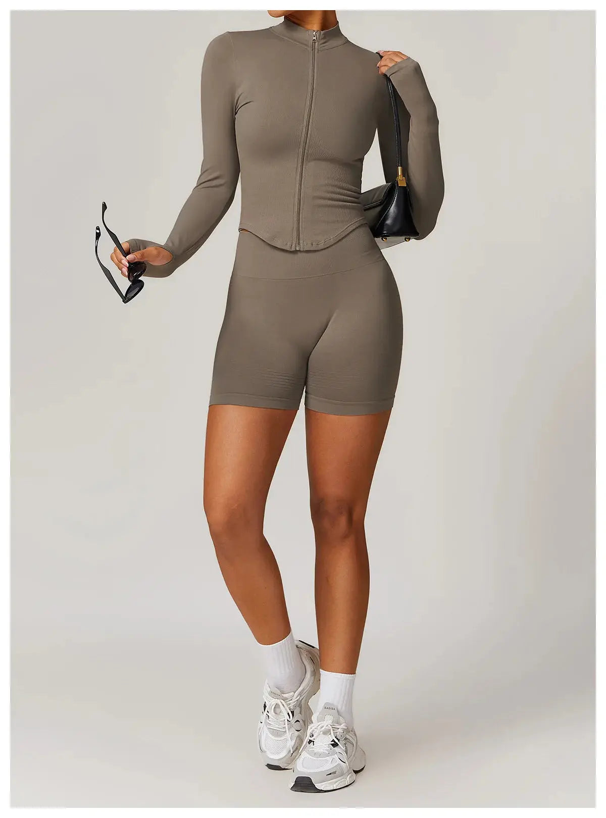 A person wearing the Mirèlle Workout Jacket with Zip & Quick Dry in taupe and matching biker shorts, paired with white socks, white sneakers, while holding black sunglasses and a small black purse.