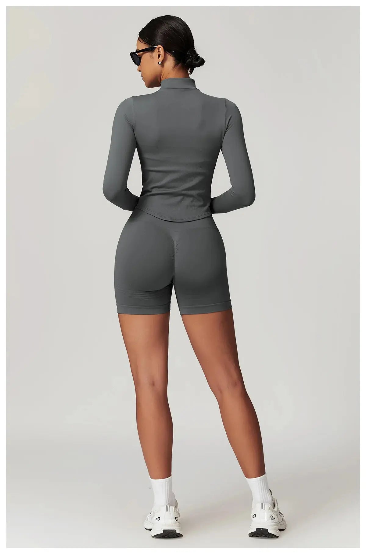 A woman stands facing away, wearing the Mirèlle Workout Jacket with Zip & Quick Dry in gray, matching shorts, white socks and sneakers, plus black sunglasses, set against a plain light background.