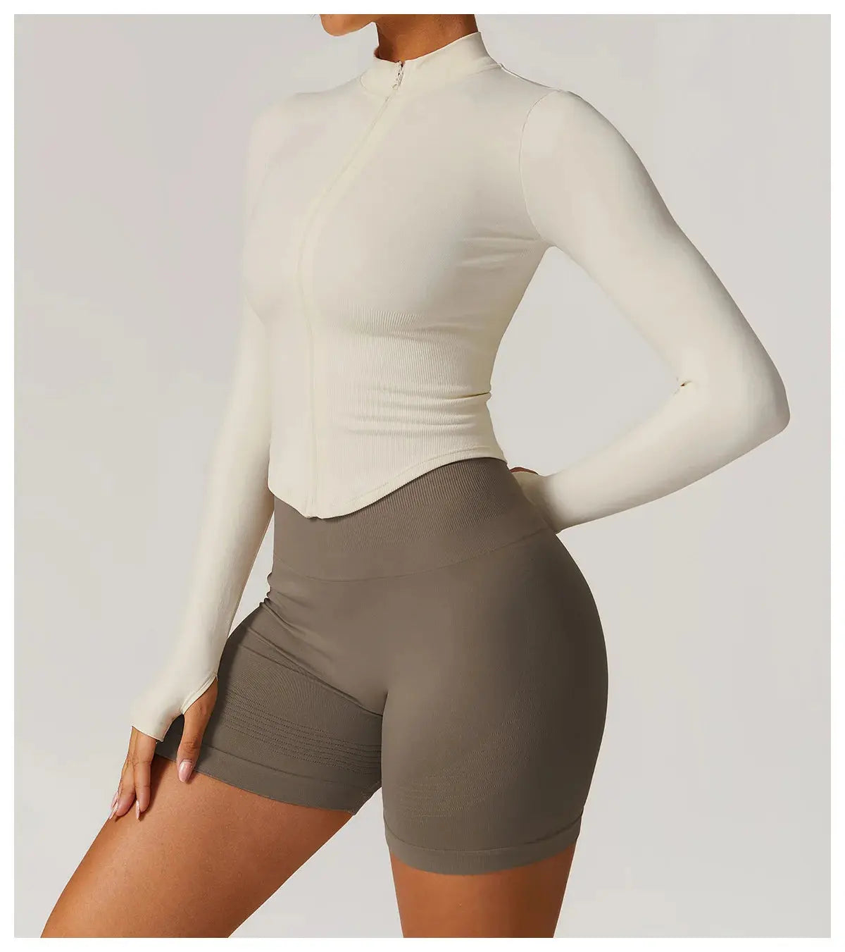 A woman poses with one hand on her hip in a Mirèlle Workout Jacket with Zip & Quick Dry in cream and high-waisted taupe bike shorts, set against a plain light background.