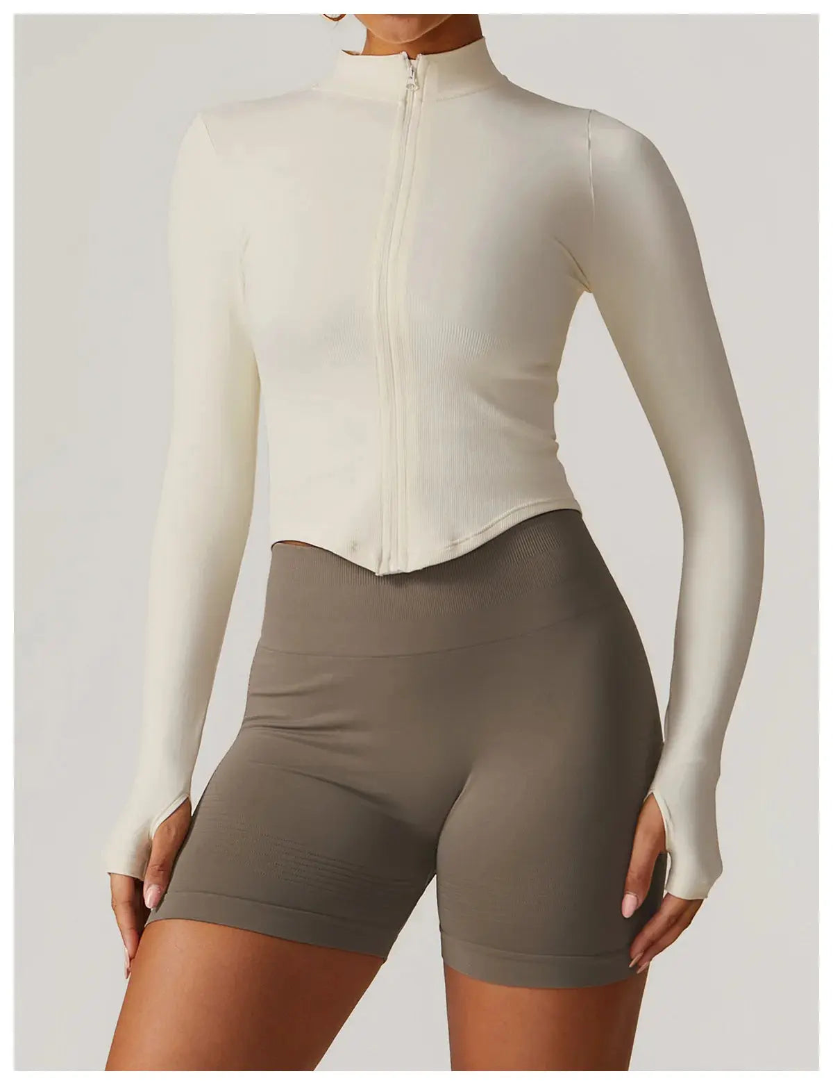 A person in a Mirèlle Workout Jacket with Zip & Quick Dry, featuring long sleeves, thumb holes, and sweat-wicking fabric, paired with high-waisted taupe biker shorts, stands against a neutral background.