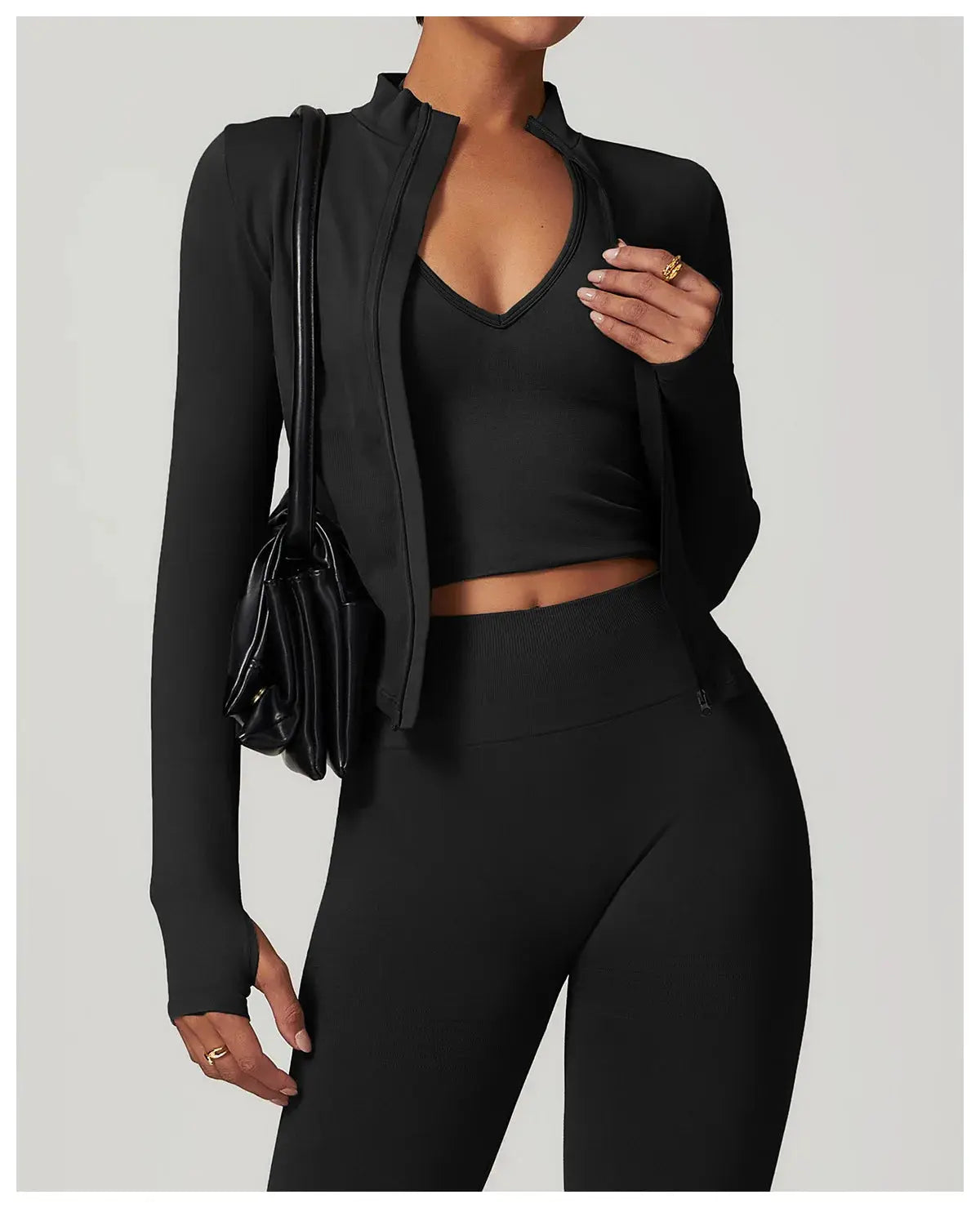A person in a black Mirèlle Workout Jacket with Zip & Quick Dry, crop top, high-waisted leggings, and a black shoulder bag poses for a photo; their face isnt visible.