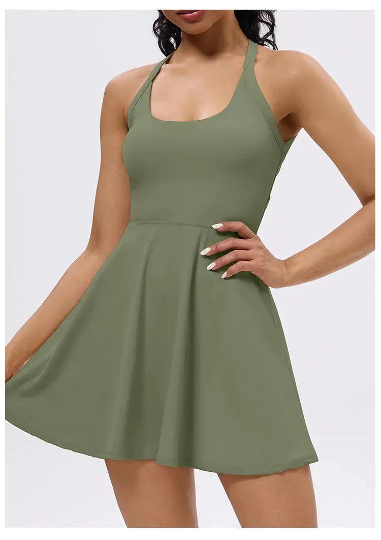 A woman models the Mirèlle Tennis Dress with Back Twist, a sleeveless olive green piece featuring a flared skirt, twist-back design, and built-in shorts with pockets. She poses against a light background, one hand on her hip.