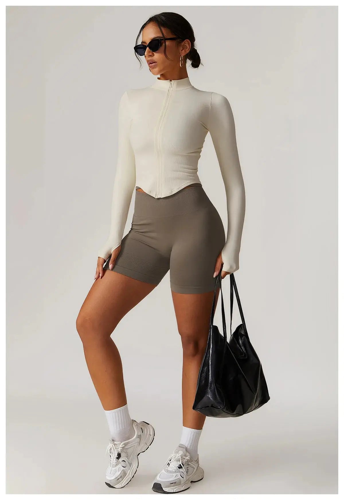 A woman in sunglasses poses against a plain light background, wearing the Mirèlle Workout Jacket with Zip & Quick Dry—a fitted, long-sleeve cream top—paired with taupe shorts, white sneakers and socks, and carrying a large black bag.