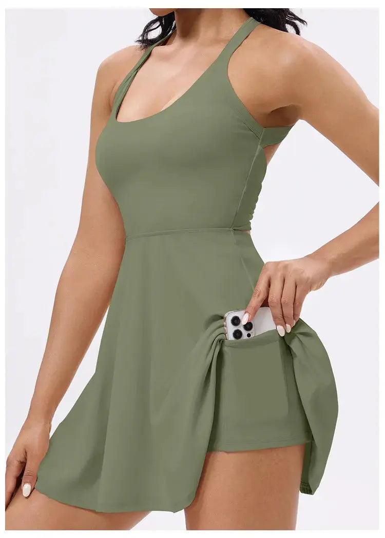 A woman wears the Mirèlle Tennis Dress with Back Twist, an olive green sleeveless dress featuring built-in shorts with pockets, her phone partially tucked into a discreet side pocket.