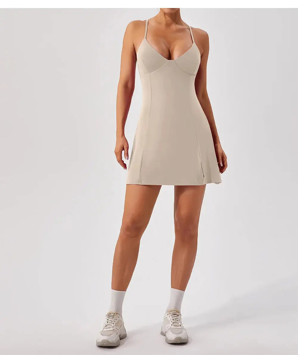 A woman stands against a plain background wearing the Mirèlle Tennis Dress Triangle Top in light beige, featuring spaghetti straps, built-in shorts, and paired with white sneakers and ankle socks for a sporty look.
